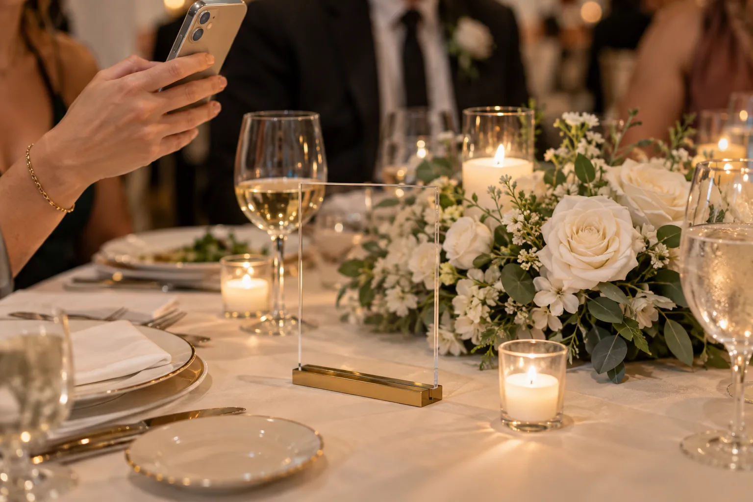 A blank acrylic sign on a gold stand beside a centerpiece, a guest holding a phone above the table