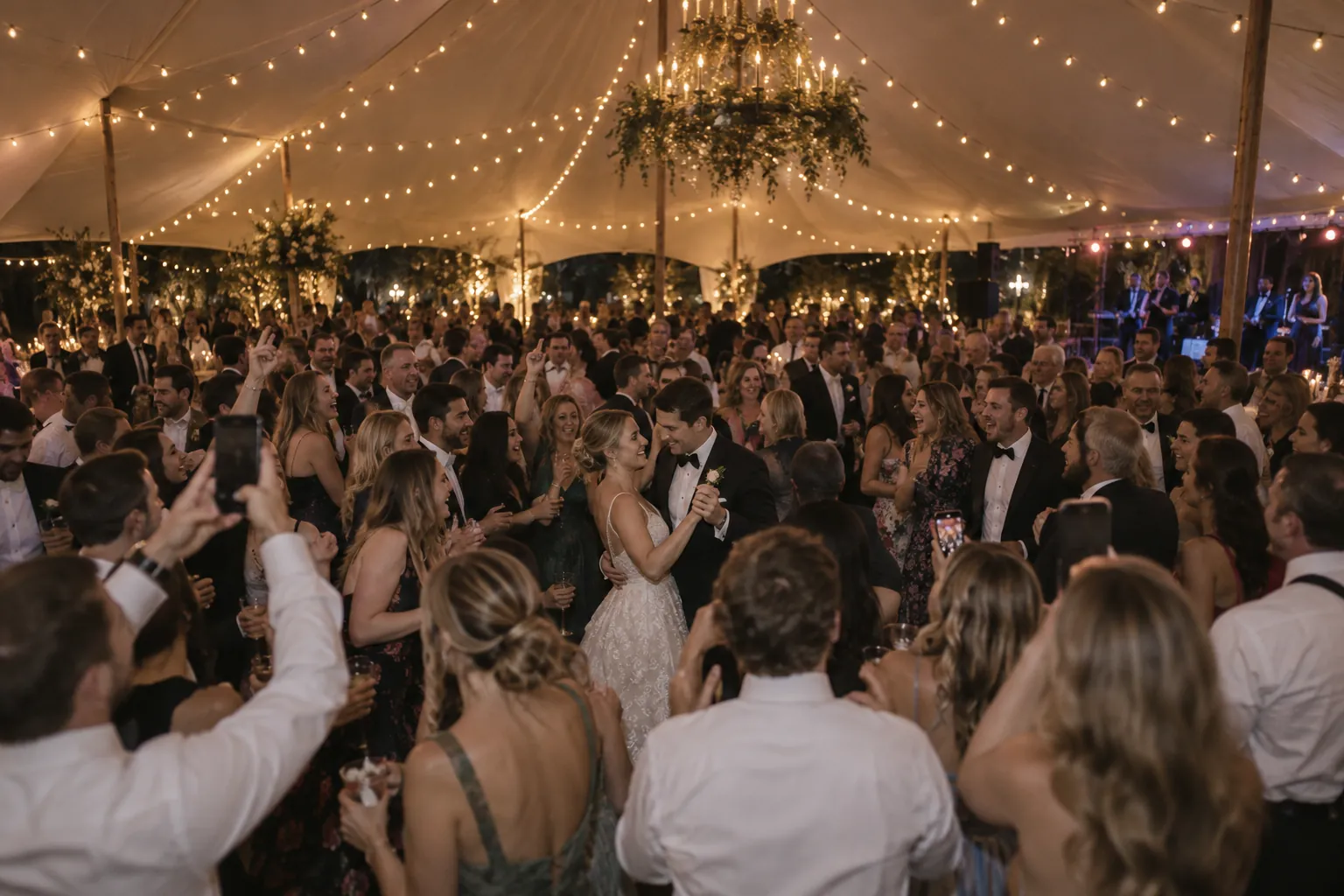 A packed dance floor under a big white tent at a large wedding, guests filming on their phones