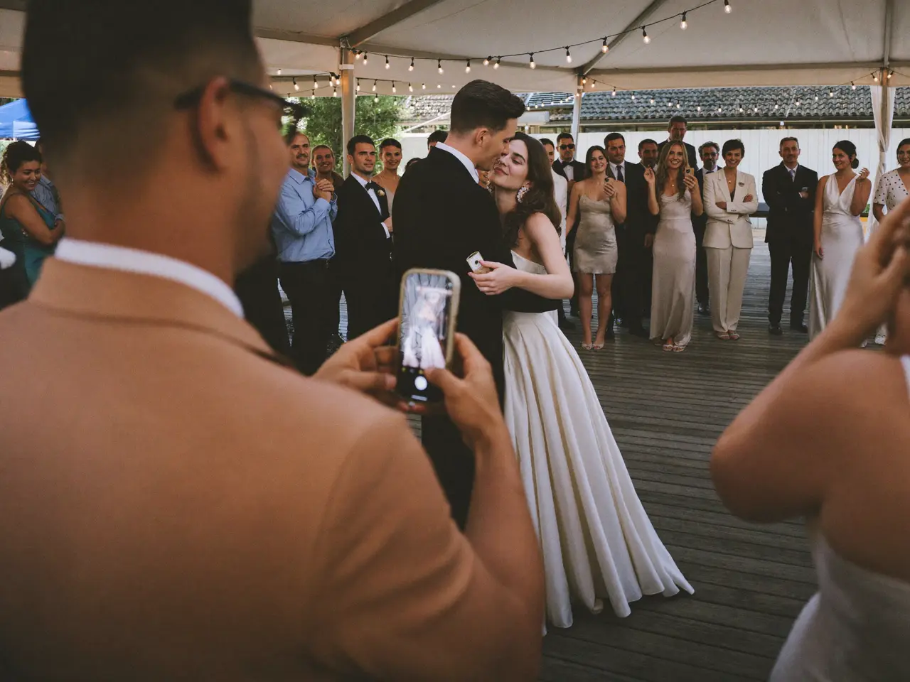 A guest in a tan suit filming the first dance on his phone while the couple dances under string lights